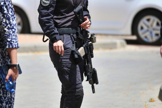 Israelischer Soldat steht im Vordergrund; Hintergrund zeigt Zerstörung und militärische Aktivitäten in Gaza.