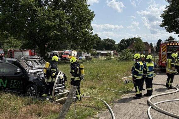 Feuerwehr löscht brennendes Fahrzeug unter Carport; Einsatzkräfte sichern angrenzendes Gebäude.
