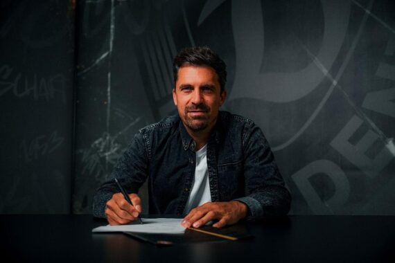 Thomas Stamm unterschreibt Vertrag, lächelt glücklich, im Hintergrund Dynamo Dresden-Logo.