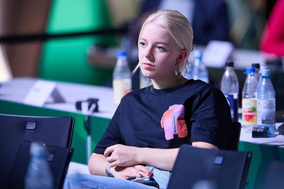 Junge Politikerin mit provokantem Pullover, umgeben von kontroversen Diskussionen in der Partei.