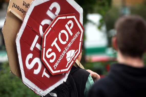 Protestierende Menschen im Hintergrund, Plakate gegen Zwangsräumungen, urbane Kulisse, emotionale Stimmung.