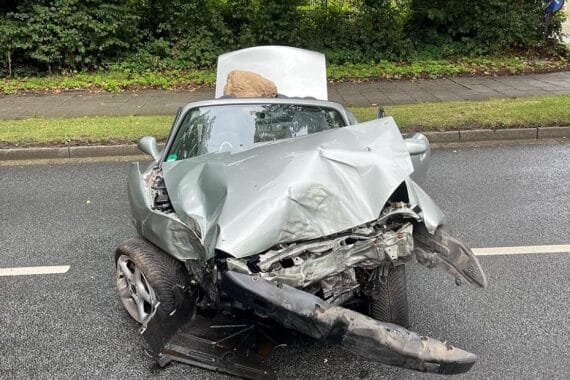 Unfallwagen auf Straße, umgeben von Polizei, Rettungswagen und beschädigten Bäumen im Hintergrund.