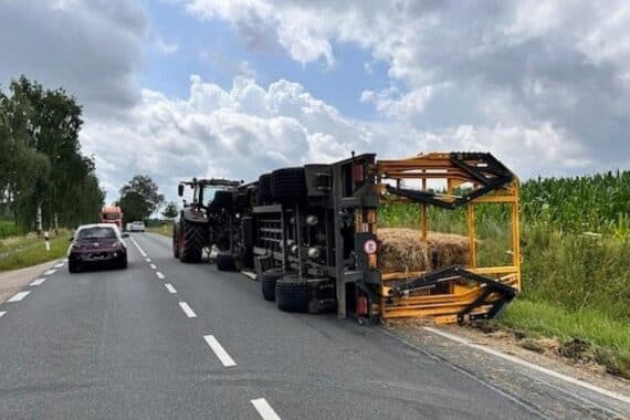 Umgekipptes Traktorgespann mit verstreuten Heuballen auf Landesstraße, Unfallstelle. Polizei sichert Beweise.