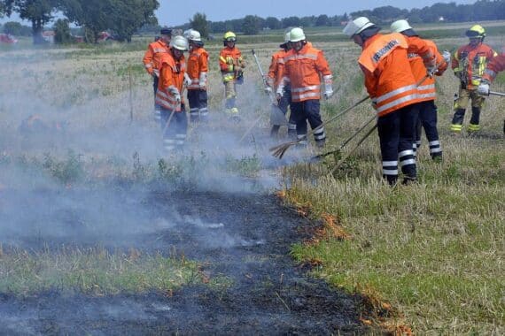 Freiwillige Feuerwehr übt Vegetationsbrandbekämpfung mit Flammen und Einsatzfahrzeugen auf einem Stoppelfeld.