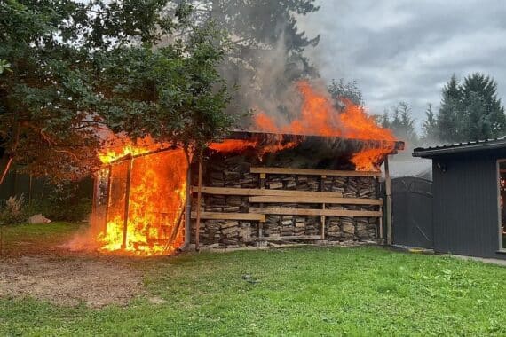 Brennendes Holzlager unter Kontrolle, Feuerwehr im Einsatz, gefahrlose Umgebung, Brandursache wird ermittelt.