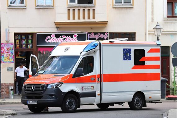 Rettungswagen am Unfallort, schwere Kollision mit zwei Autos, Straßenlaterne beschädigt, Einsatzkräfte tätig.