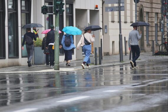 Personen mit Regenschirmen unter wechselhaftem Wetter, Sturmwolken und Sonnenschein.