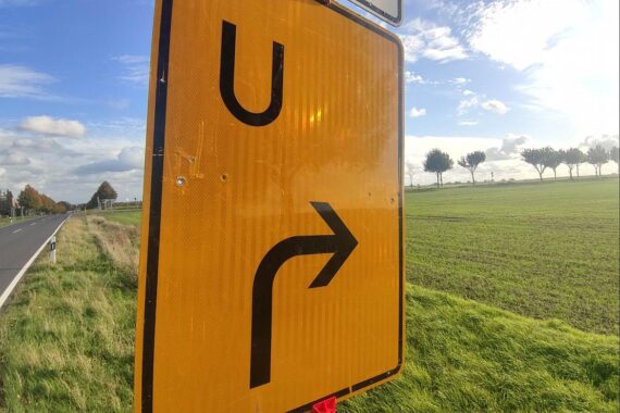 Umleitungsschild mit Baustellenhinweisen und Verkehrsteilnehmer, die Geduld fordern.