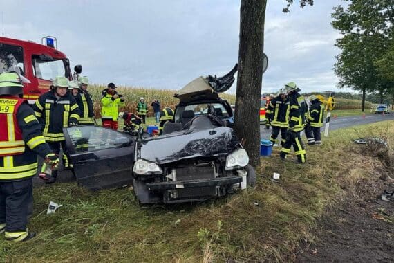 Zerstörtes Fahrzeug prallt gegen Baum, Unfallstelle auf regennasser Fahrbahn, Rettungskräfte im Einsatz.
