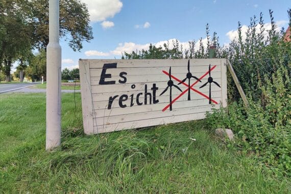 Ministerpräsidenten diskutieren Windkraft, während Bürgerprotest gegen neue Windräder im Hintergrund sichtbar ist.