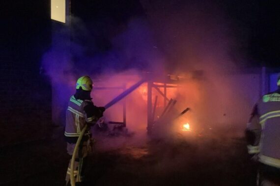 Flammendes Carport neben Wohnhaus, Feuerwehr im Einsatz, rauchende Szenerie, Gefahr für Nachbarn.