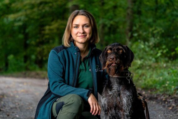 Lia Ternes steht im Wald, lächelnd, mit Forstwerkzeugen. Symbolisiert ihr neues Amt als Forstrevierleiterin.