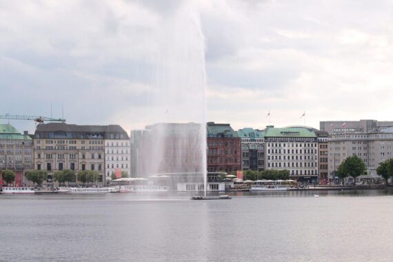 Wechselhaftes Wetter über Hamburg mit Wolken, Regen und der Binnenalster-Fontäne im Vordergrund.