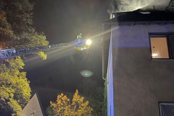 Feuerwehr im Einsatz, evakuierte Bewohner, rauchendes Mehrfamilienhaus, Einsatzkräfte prüfen Dach.