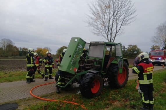 Ein Traktor brennt am Straßenrand; Feuerwehrleute löschen den Brand mit Atemschutz.