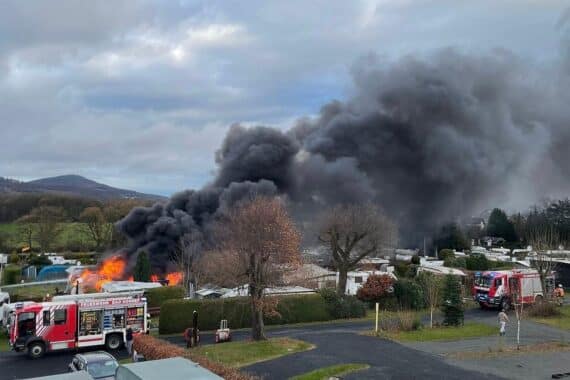 Zwei brennende Campingwagen mit starkem Rauch, Feuerwehr im Einsatz, dramatische Situation am Campingplatz.