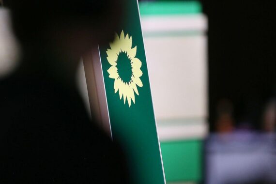 Grünen-Logo vor einem Hintergrund von vandalisierten Häusern und Autos, Symbol für Gewalt und Einschüchterung.