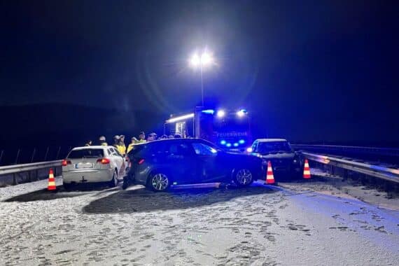 Schneebedeckte Autobahn, verunfallte Fahrzeuge, Polizeiauto, Feuerwehr und verletzte Personen am Unfallort.
