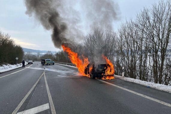 PKW in Flammen auf einer Landstraße, Feuerwehr löscht Brand, rauchende Trümmer.