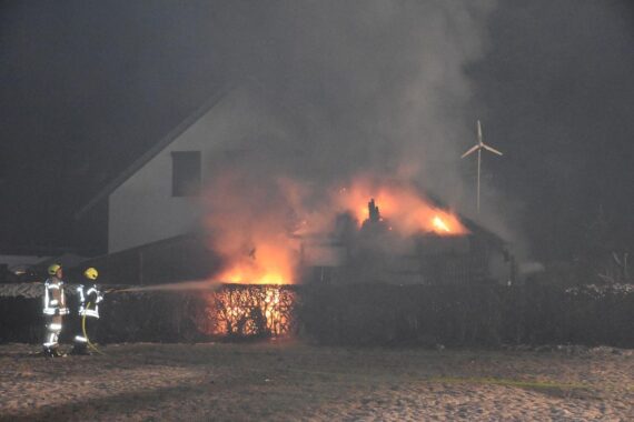 Ein abgebrannter Carport mit Feuerwehr im Einsatz, angrenzendes Wohnhaus sichtbar.