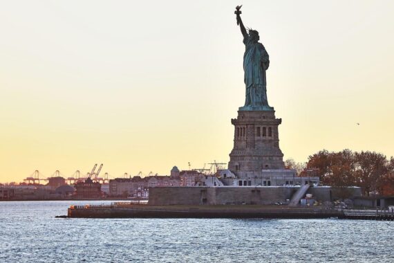 Industrieanlagen und Hafen mit Freiheitsstatue, symbolisieren Handelskonflikt und wirtschaftliche Unsicherheit.