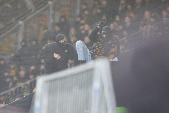 Aggressive, vermummte Fußballfans während Ausschreitungen im Stadion, Polizei im Hintergrund.