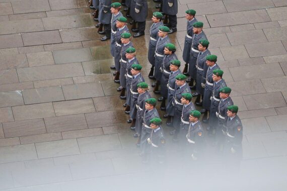 Soldaten der Bundeswehr stehen im Vordergrund, im Hintergrund NATO-Symbole und militärische Ausrüstung.