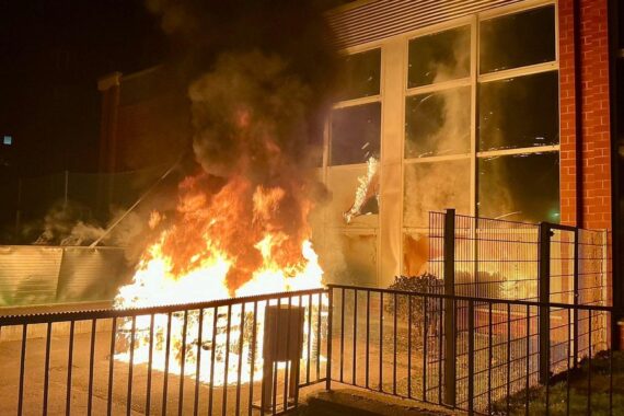 Feuerwehr im Einsatz bei Brand nahe Sporthalle, Flammen und Rauch sichtbar.