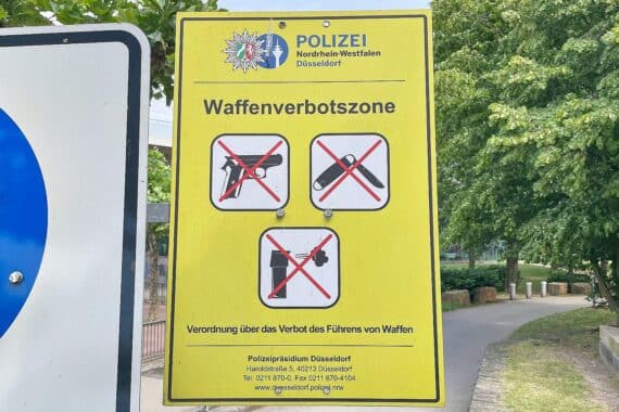 Schild einer Waffenverbotszone in Kiel mit Polizeipräsenz und Stadtansicht im Hintergrund.
