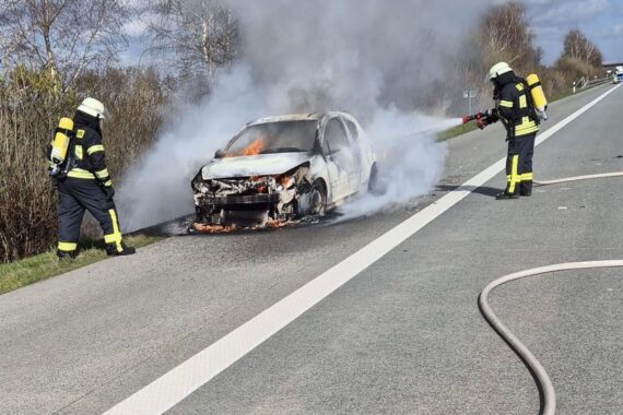 Feuerwehr löscht brennenden Kleinwagen auf Autobahn, Rauchentwicklungen beeinträchtigen Sicht.