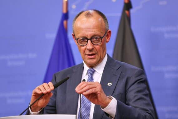 Friedrich Merz am Telefon mit ernstem Gesichtsausdruck, Hintergrund könnten politische Symbole oder Flaggen sein.