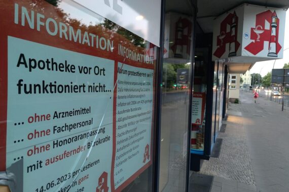 Apothekerprotest vor einer Apotheke, Transparent mit Forderungen, besorgte Bürger im Hintergrund.