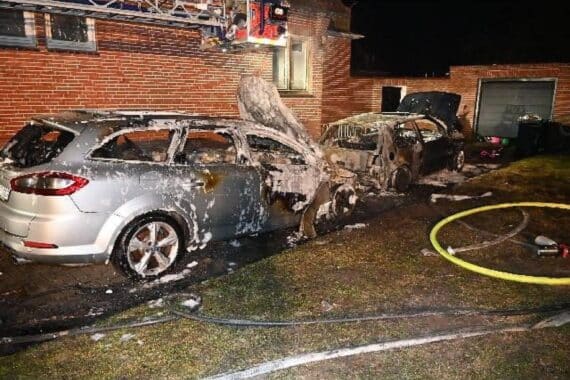 Zwei komplett verbrannte Autos vor einem Mehrfamilienhaus, Feuerwehr im Einsatz, Schadensursache unklar.