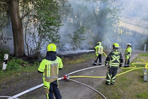 Löschende Feuerwehrmänner bekämpfen einen Waldbrand, dichte Rauchentwicklung über brennendem Unterholz sichtbar.