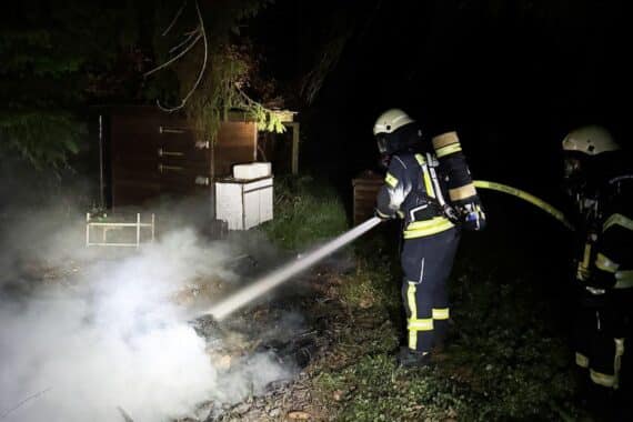 Feuerwehr bekämpft Flächenbrand im Wald, schützt Gartenhütten vor weiterer Ausbreitung.