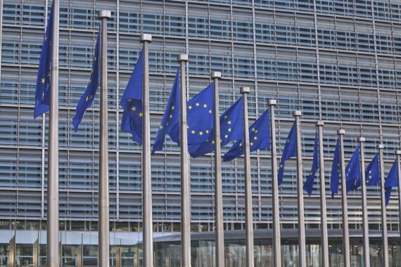 EU-Fahnen im Vordergrund, Politikdebatte über Einstimmigkeitsprinzip, symbolisiert durch neutralen Hintergrund und gerissene Grenzen.