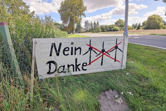 Protestierende Menschen gegen Windräder, Transparente, naturnahe Umgebung, Bäume und Windkraftanlagen im Hintergrund.