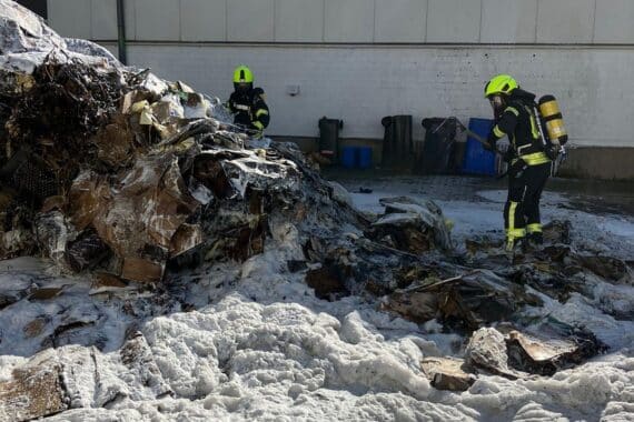 Eine brennende Müllpresse im Innenhof eines Gewerbebetriebs mit Feuerwehr im Einsatz.