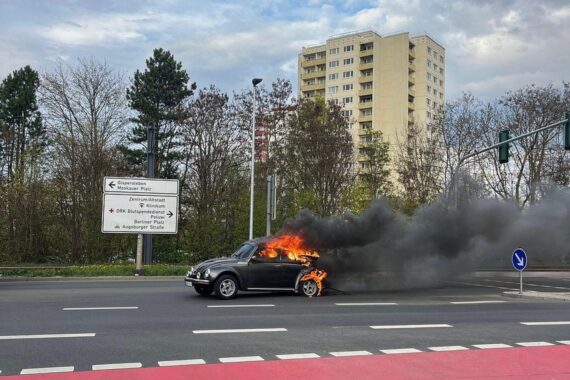 Brennender VW Käfer auf der Straße, Feuerwehr im Einsatz, Rauch und Zerstörung.