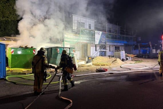 Feuerwehr löscht brennenden Baucontainer, Flammen drohen auf angrenzendes Garagendach überzugreifen.