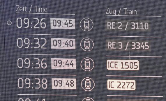 Anzeigetafel an einem Bahnhof: Gespräch über verspätete Fernzüge, Bahnprobleme und Pünktlichkeitsziel bis 2029.