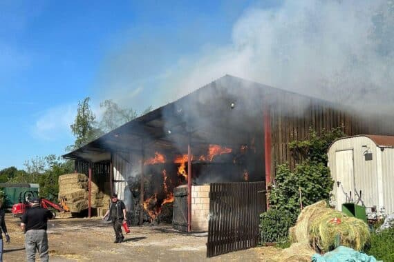 Brand einer Lagerhalle mit Heuballen, Feuerwehr im Einsatz, dichte Rauchentwicklung, ländliche Umgebung.
