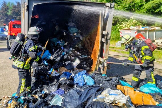 Löscharbeiten an Müllpresse, Feuerwehr im Einsatz, Rauchentwicklung, Feuerwehrfahrzeuge, Klinikgelände.