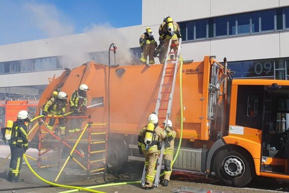 Rauchender Müllwagen, Feuerwehr bei Löscharbeiten, Hinweis auf unsachgemäße Müllentsorgung und Brandgefahr.