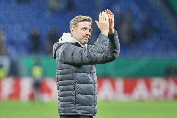 Florian Kohfeldt lächelt, trägt Trainer-Outfit und steht stolz im Stadion von Darmstadt 98.