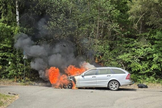 Ein ausgebrannter Mercedes steht an der Straße, Feuerwehr löscht den Brand.