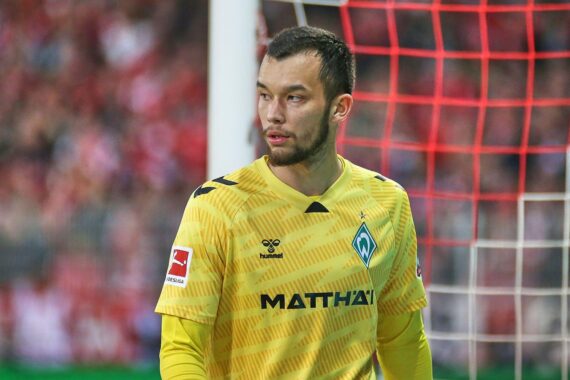 Mio Backhaus in SC Freiburg-Trikot, Torwarthandschuhe, entschlossenes Gesicht, Stadionhintergrund, sportlicher Fokus.