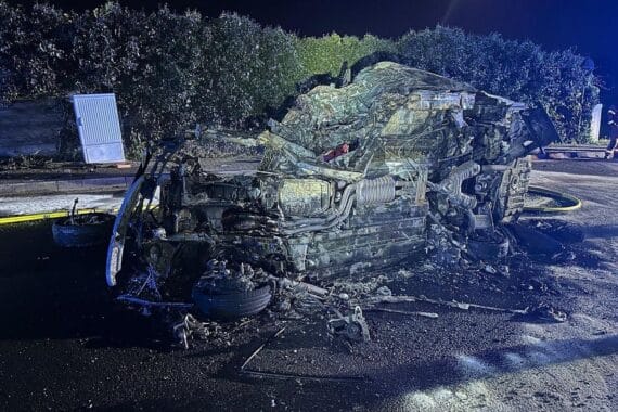 Schwerer Verkehrsunfall: brennendes Auto, Einsatzkräfte, verletzter Fahrer, gesperrte Straße.