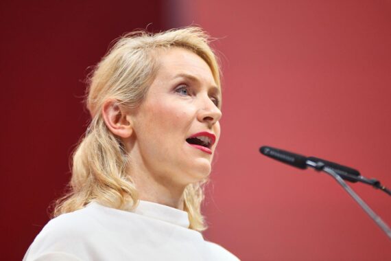 Ines Schwerdtner kritisiert Wohngeldkürzungen der Regierung, hinter ihr ein symbolisches Bild von Unsicherheit.