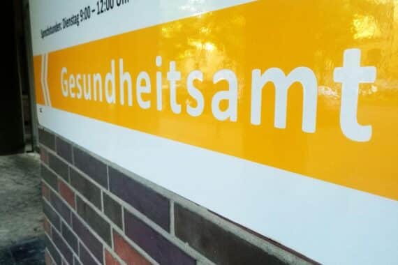 Gesundheitsamt-Mitarbeiter in einer Besprechung, diverse Personen, Akten und Laptops sichtbar.